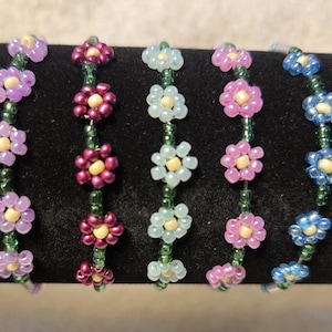 May include: Five beaded bracelets with flower designs in various colours, including purple, magenta, light blue, and green. Each bracelet features small, colourful beads arranged to resemble flowers, strung on a green thread.