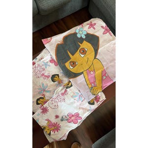Puede incluir: Un juego de sábanas con el tema de Dora la Exploradora. La funda de almohada presenta una imagen grande de Dora con una flor en el pelo. Las sábanas tienen un patrón repetido de Dora, flores y estrellas en rosa, azul y amarillo.