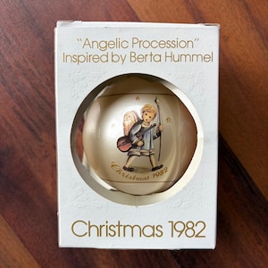 Può includere: Una palla di Natale in una scatola. L'ornamento sferico presenta un'immagine dipinta di un angelo che suona uno strumento a corde. La scatola recita "Angelic Procession" e "Christmas 1982". L'ornamento è in una scatola bianca con una finestra circolare.