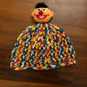 May include: A knitted beanie hat in a vibrant mix of colours, including blue, yellow, red, and white. A plush character head is attached to the hat. The character has an orange face, black hair, and a red nose.