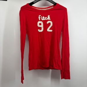 May include: A long-sleeved red top featuring the white text fitch and the number 92 in white, textured, raised lettering on the front. The garment has a crew neckline and is displayed on a black hanger against a plain white background.