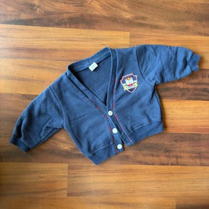 May include: A navy blue vintage cardigan with a V-neck and long sleeves. The cardigan has white buttons and a red and white trim along the button line. A patch with a bear and the word "Campus" is on the upper left chest.