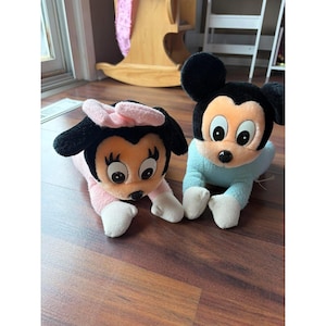 May include: Two plush toys featuring cartoon characters with large eyes and black ears. One character has a peach-colored face, a pink bow, and a pink body. The other character has a peach-colored face, a black head, and a light blue body.