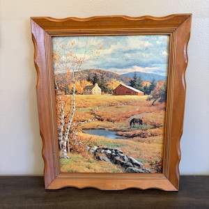 May include: A framed landscape painting depicting an autumn scene. The artwork features a red barn, a white house, a horse grazing, and a stream. The frame is made of light brown wood with a decorative edge.