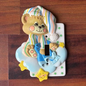 Puede incluir: Una cubierta decorativa para interruptor de luz con un oso de dibujos animados con gorro de dormir y pijama a rayas, descansando sobre una nube. La cubierta tiene un fondo blanco con estrellas verdes y acentos de estrella amarilla. La abertura del interruptor es negra.