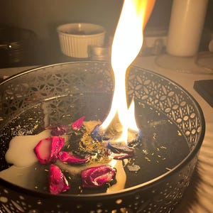 Puede incluir: Una vela encendida en un cuenco decorativo negro. La vela tiene una gran llama y está rodeada de cera derretida, hierbas secas y pétalos de rosa rojos. El cuenco tiene un diseño decorativo calado.