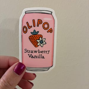 May include: A die-cut sticker shaped like a soda can with a light pink background. The text OLIPOP is printed in orange at the top, with a graphic of a strawberry and a white flower in the center. The words Strawberry Vanilla are printed in black below.