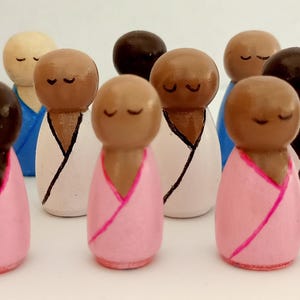 Op de afbeelding: Een verzameling handbeschilderde houten peg dolls. Sommige poppen zijn beschilderd met lichte huidtinten, andere met donkere huidtinten. De poppen dragen gewaden in blauwe, witte en roze tinten. Elke pop heeft gesloten ogen.