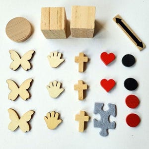 Puede incluir: Un conjunto de suministros de artesanía de madera que incluye dos bloques de madera, un disco redondo de madera, una flecha de madera, tres mariposas de madera, tres manos de madera, tres cruces de madera, tres corazones rojos, tres círculos negros, una pieza de rompecabezas gris y dos círculos rojos.