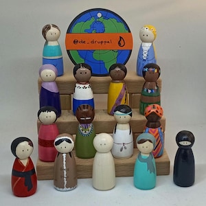 Op de afbeelding: Een set van 15 houten poppen die verschillende culturen en etniciteiten vertegenwoordigen. De poppen zijn geschilderd in verschillende kleuren en ontwerpen, waaronder rood, blauw, groen, geel en zwart. De poppen zijn in een verspringende formatie op een houten plank geplaatst. Een globe met de tekst "@de_druppel" is boven de poppen geplaatst.