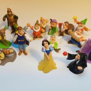 Op de afbeelding: Een verzameling van 14 plastic figuurtjes uit het sprookje Sneeuwwitje en de Zeven Dwergen. De figuurtjes omvatten Sneeuwwitje, de boze koningin, Prins Charming, de Zeven Dwergen en een hert. De figuurtjes staan allemaal in verschillende poses en zijn geschilderd in felle kleuren.