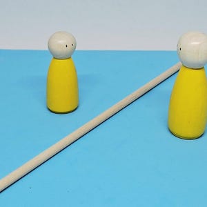 Può includere: Due bambole di legno gialle con teste bianche e occhi a puntini neri sono su una superficie blu, con un lungo tassello di legno tra loro. Le bambole hanno un design semplice e minimalista.