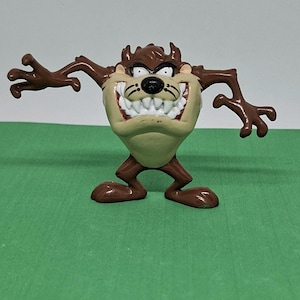 Op de afbeelding: Een bruin en tan Taz-beeldje met een brede grijns en uitgestrekte armen. Het stripfiguur heeft een tan gezicht, witte tanden en bruine vacht. Het beeldje staat op een groene ondergrond.