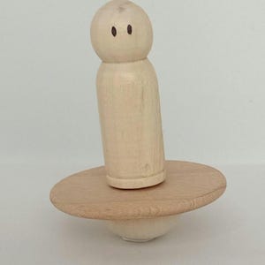 Könnte beinhalten: Ein Holzspielzeug mit einer runden Basis und einer kleinen Holzfigur mit einem gemalten Gesicht. Die Figur ist auf der Basis balanciert.