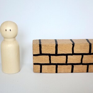 Op de afbeelding: Een houten peg doll met een afgeronde kop en twee kleine zwarte stippen voor ogen staat naast een rechthoekige houten doos. De doos is beschilderd om op een bakstenen muur met zwarte lijnen te lijken.