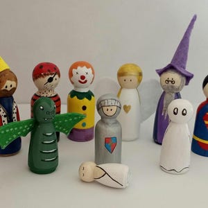 Op de afbeelding: Een verzameling handgeschilderde houten peg dolls die verschillende personages uitbeelden. De set bevat een koning, piraat, clown, draak, ridder, engel, baby, geest, tovenaar en superheld. Elke pop is uniek ontworpen.