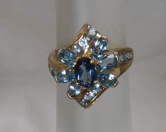Puede incluir: Un anillo de oro con una gran piedra ovalada azul rodeada de piedras azules más pequeñas y piedras transparentes. El anillo está engastado en un diseño floral.