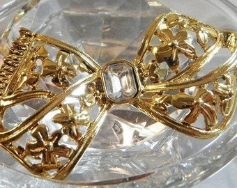 May include: A gold-tone bow with a clear crystal center, set on a clear glass base. The bow is intricately detailed with floral designs.