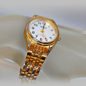 May include: A gold-tone wristwatch with a white face and black hands. The watch has a gold-tone metal band and says "Berus Quartz 100m Water Resist Alarm" on the face.