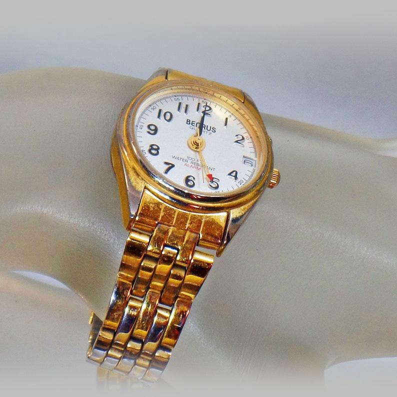 May include: A gold-tone wristwatch with a white face and black hands. The watch has a gold-tone metal band and says "Berus Quartz 100m Water Resist Alarm" on the face.