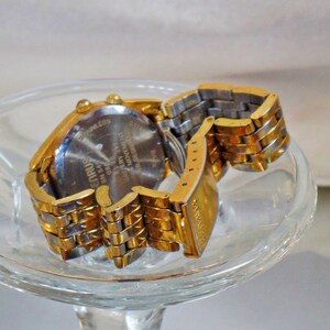 May include: A gold-tone wristwatch with a stainless steel band. The watch face is silver with black markings. The back of the watch has the words "Stainless Steel" and "Water Resistant" printed on it. The watch is on a clear glass surface.