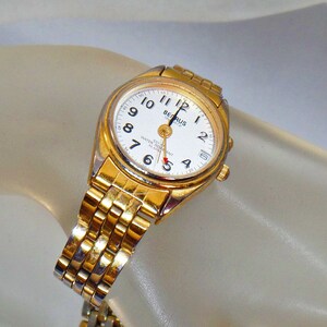 May include: A gold-toned wristwatch with a white face and black hands. The watch has a gold-toned metal band and says "BENRUS QUARTZ" on the face. The watch is water resistant and has an alarm.