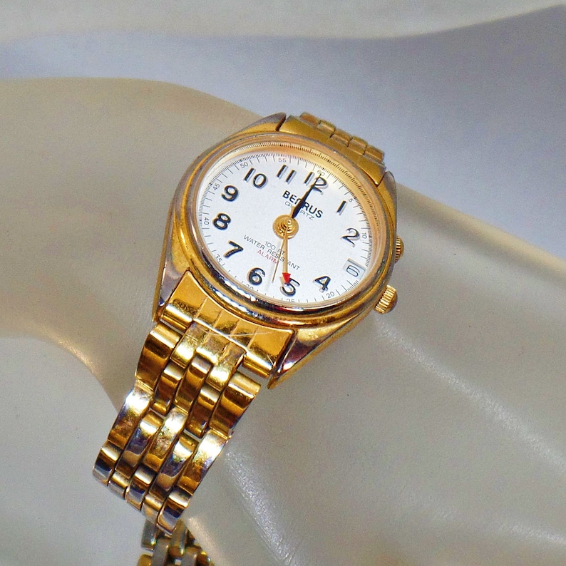May include: A gold-toned wristwatch with a white face and black hands. The watch has a gold-toned metal band and says "BENRUS QUARTZ" on the face. The watch is water resistant and has an alarm.
