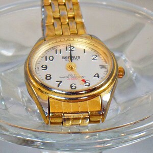 May include: A gold-tone wristwatch with a white face and black hands. The watch has a gold-tone metal band and a date window at the 3 o'clock position. The watch face also has the text "BENRUS QUARTZ" and "100 F WATER RESISTANT ALARM".