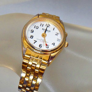 May include: A gold-tone wristwatch with a white face and black hands. The watch has a gold-tone metal band and says "BENRUS QUARTZ" and "100 FT WATER RESISTANT ALARM" on the face.