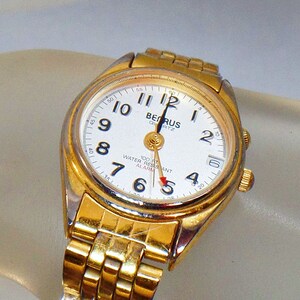May include: A gold-tone wristwatch with a white face and black hands. The watch has a date window and an alarm function. The watch is water resistant and has a quartz movement. The text "BERUS QUARTZ" is printed on the face of the watch.