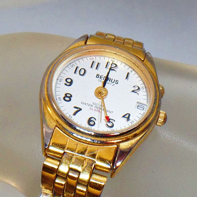 May include: A gold-tone wristwatch with a white face and black hands. The watch has a date window and an alarm function. The watch is water resistant and has a quartz movement. The text "BERUS QUARTZ" is printed on the face of the watch.