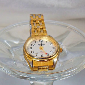 May include: A gold-tone wristwatch with a white face and black hands. The watch has a gold-tone band and a silver bezel. The face of the watch has the words "BENRUS QUARTZ" and "100 FT WATER RESISTANT ALARM" printed on it.