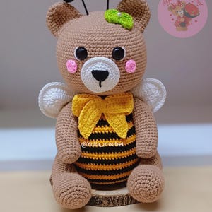 May include: A handmade crochet bee-themed bear. The bear is primarily brown with a yellow and black striped belly, a yellow bow, and white wings. It has black eyes, pink cheeks, and a green bow on its head. The bear is sitting on a wooden slice.