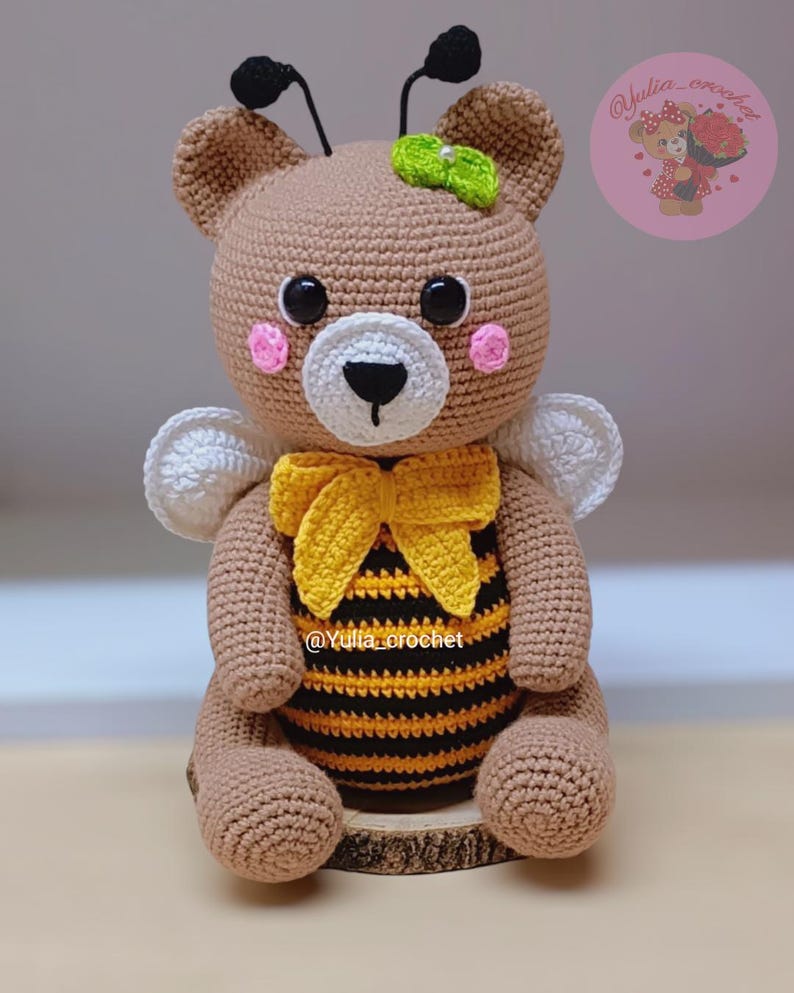 May include: A handmade crochet bee-themed bear. The bear is primarily brown with a yellow and black striped belly, a yellow bow, and white wings. It has black eyes, pink cheeks, and a green bow on its head. The bear is sitting on a wooden slice.