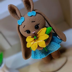 May include: A crocheted brown bunny doll with blue bows, wearing a blue dress, and holding a yellow sunflower. The doll has large black eyes and a pink nose. The text "@Yulia_crochet" is visible on the flower.