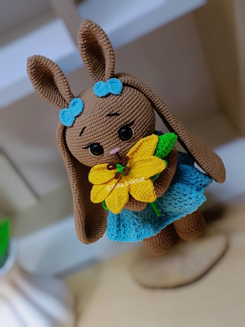 May include: A crocheted brown bunny doll with blue bows, wearing a blue dress, and holding a yellow sunflower. The doll has large black eyes and a pink nose. The text "@Yulia_crochet" is visible on the flower.