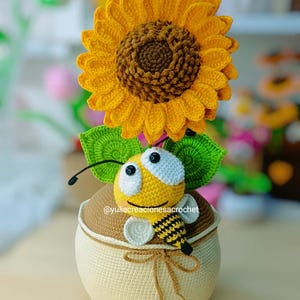 Puede incluir: Un girasol de ganchillo hecho a mano, con un centro marrón y pétalos amarillos, se encuentra sobre un recipiente beige y marrón. Una abeja amarilla y negra con ojos grandes está posada en el recipiente, con hojas verdes. El texto "@yuliacreacionesacrochet" es visible.