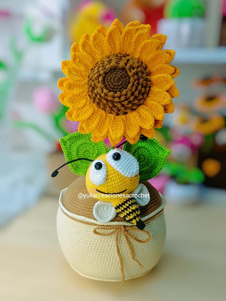 May include: A handmade crochet sunflower with a brown centre and yellow petals sits on a beige and brown container. A yellow and black bee with large eyes is perched on the container, with green leaves. The text "@yuliacreacionesacrochet" is visible.