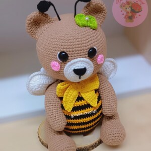 May include: A handmade crochet teddy bear with bee-themed details. The bear is brown with a white muzzle, pink cheeks, and black eyes. It has a yellow and black striped belly, a yellow bow, and white wings. Black antennae and a green leaf adorn the head.