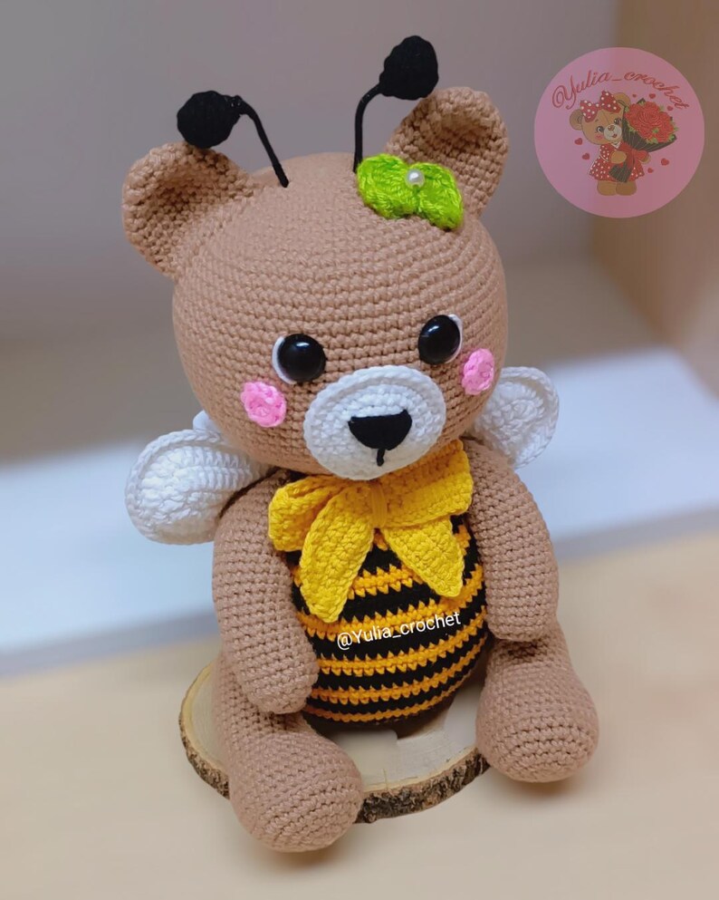 May include: A handmade crochet teddy bear with bee-themed details. The bear is brown with a white muzzle, pink cheeks, and black eyes. It has a yellow and black striped belly, a yellow bow, and white wings. Black antennae and a green leaf adorn the head.