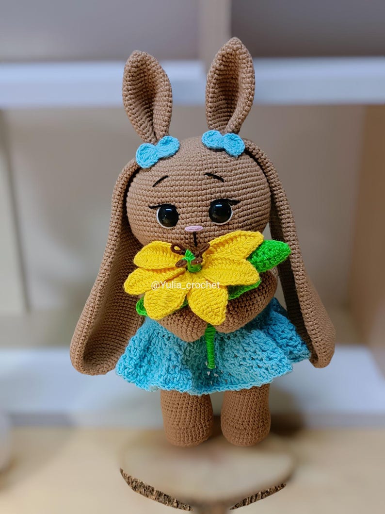 May include: A crocheted bunny doll in shades of brown, with a blue dress and a yellow flower. The bunny has long ears, blue bows, and large black eyes. The doll is displayed on a wooden stand.
