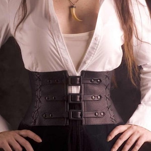 May include: A black leather corset with multiple buckles and straps. The corset is worn over a white shirt.
