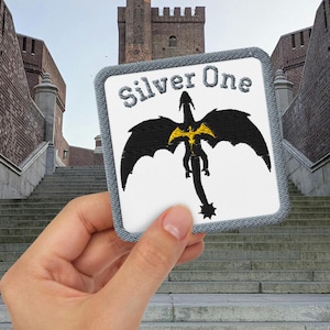 May include: A square patch featuring the text "Silver One" and a black dragon with yellow detailing. The patch is held in front of a brick building with a stone staircase.