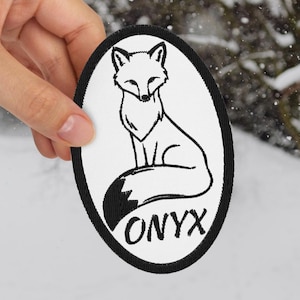 May include: An oval patch featuring a black and white fox illustration with the word "ONYX" below. The patch has a black border and is held against a snowy outdoor background.
