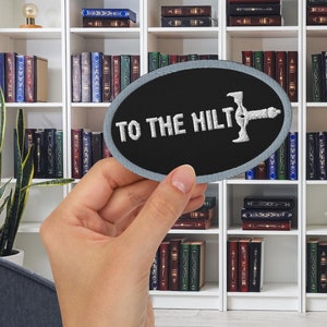 以下が含まれることがあります： 白いステッチが付いた灰色の楕円形のワッペン。ワッペンには「To the hilt」という文字と、スタイリッシュな剣のデザインが施されています。