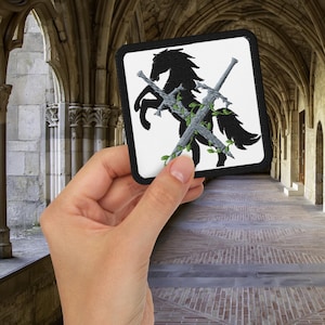 Könnte beinhalten: Ein weißer Patch mit einer schwarzen Silhouette eines Pferdes mit zwei gekreuzten Schwertern dahinter. Die Schwerter haben grüne Ranken, die darauf wachsen.