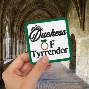 Pu&ograve; includere: Una toppa quadrata con bordo verde e le parole "Duchess of Tyrrendor" in corsivo nero. La toppa presenta una corona e un disegno ad anello. La toppa &egrave; tenuta in mano su uno sfondo di arcate in pietra.