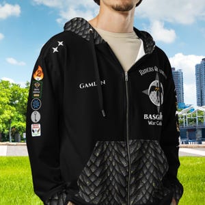 May include: Black zip-up hoodie with a quilted pattern on the lower front and hood. The hoodie features the text "GAMELN" and "Riders of the Basg War Cal" with various patches and star designs. The background is a green field and blue sky.