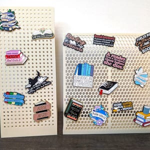 May include: Two off-white pegboards displaying enamel pins with book-themed designs. The pins feature phrases like "Book Nerd," "Reading is my Superpower," and "Drink Coffee, Buy Books, + Repeat." The pins are colorful and depict stacks of books, coffee cups, and cats.