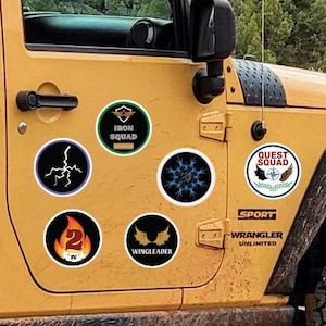 May include: A yellow Jeep Wrangler Unlimited with several round stickers on the side. A golden retriever dog is looking out the window. The stickers have various designs and text, including "Iron Squad" and "Quest Squad".